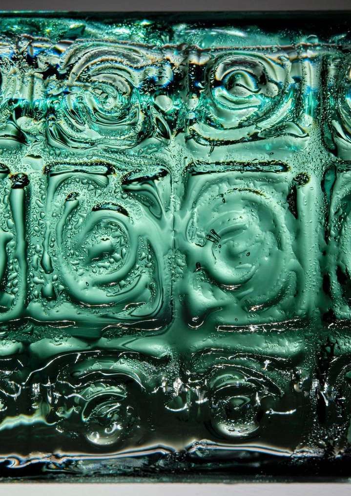 Close-up of an abstract geometric pattern in teal glass, showing wet condensation and intricate swirl designs with a shallow depth of field.
