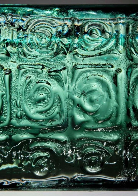 Close-up of an abstract geometric pattern in teal glass, showing wet condensation and intricate swirl designs with a shallow depth of field.