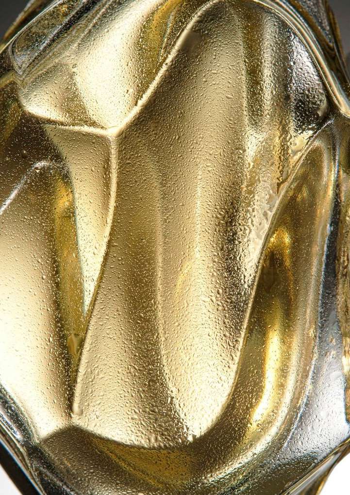 Abstract macro photograph of an ornamental gold glass vase with textured patterns and water droplets on its surface, casting soft shadows.