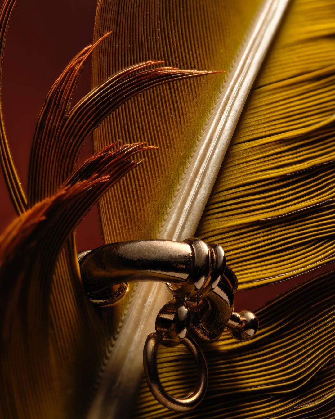 Close-up product shot of a gold metal jewelry clasp attached to brown and gold feathers on a textured brown background, indoors.