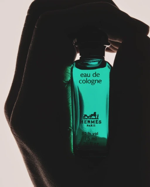 Close-up of a turquoise perfume bottle with Hermes branding, held in a person's silhouetted hands against a soft, diffused background.