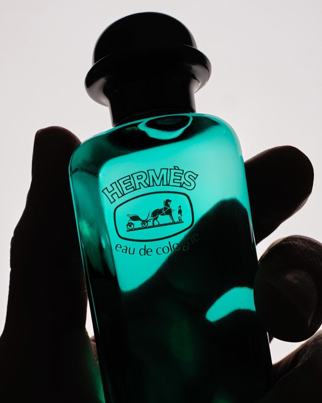 Close-up studio shot of a green-tinted glass Hermes Eau de Cologne bottle held in a hand against a white background, showcasing its branding.