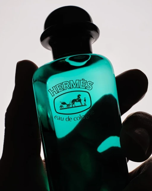 Close-up studio shot of a green-tinted glass Hermes Eau de Cologne bottle held in a hand against a white background, showcasing its branding.