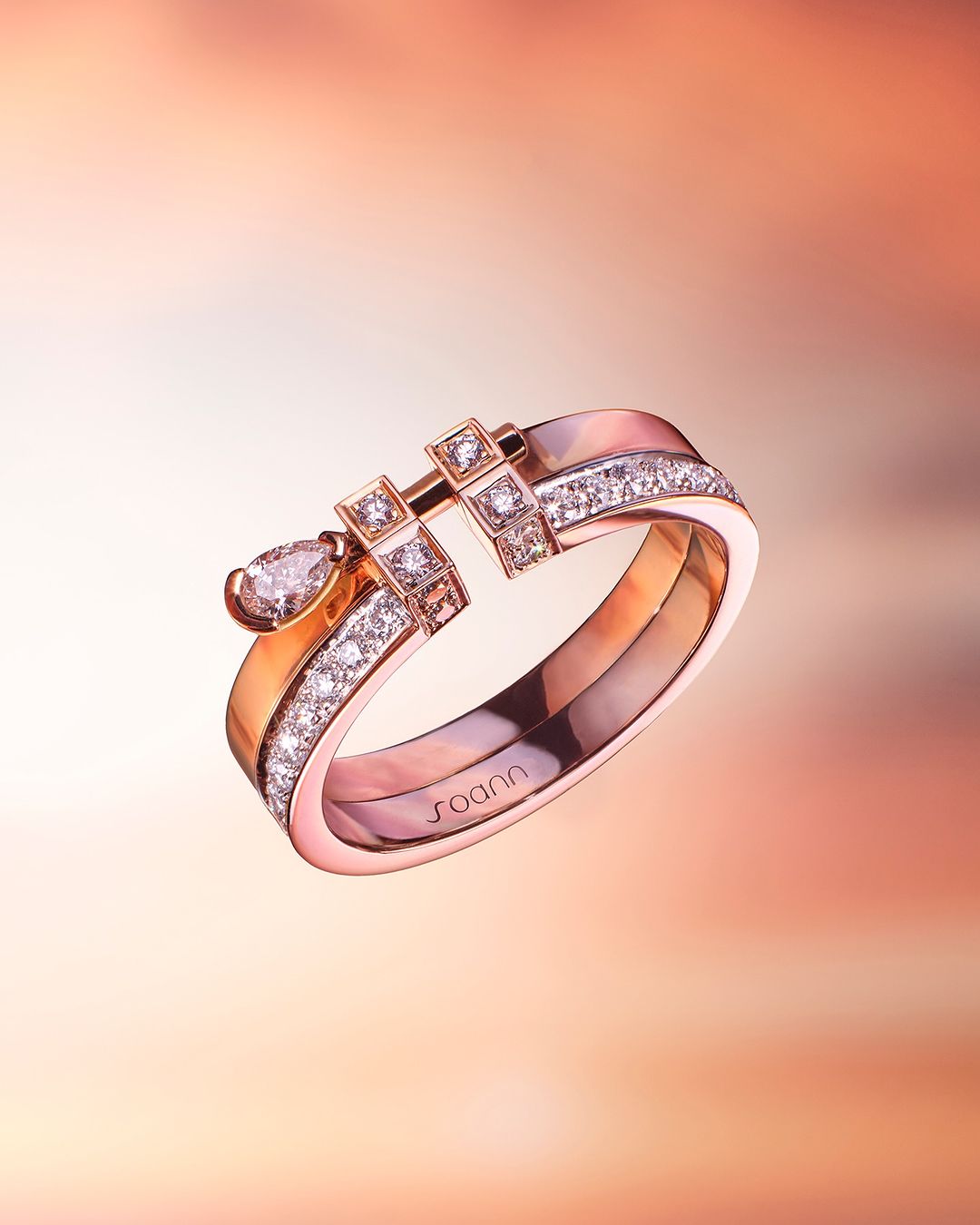 Close-up of a two-tiered gold ring featuring small, round-cut diamonds and a tear-shaped gemstone against a peach and orange gradient backdrop.