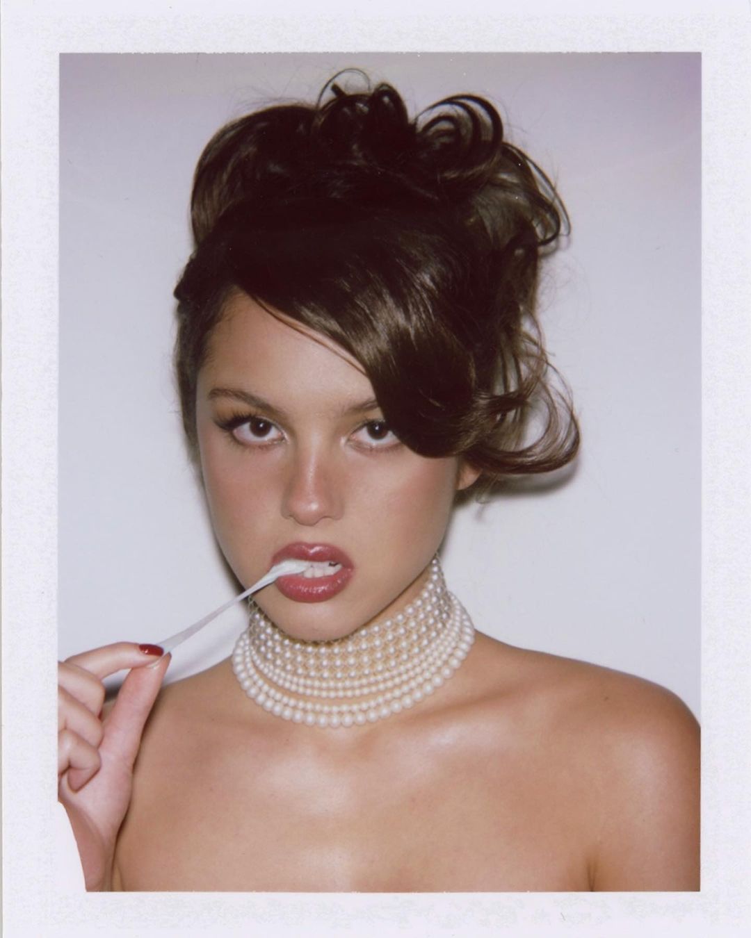 A young adult woman with dark hair and pearl necklace poses against a white background, blowing a bubble with chewing gum.