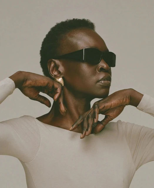 Dark-skinned woman with short hair wearing sunglasses and earrings, posing in a light-colored long-sleeved shirt in a photo studio.