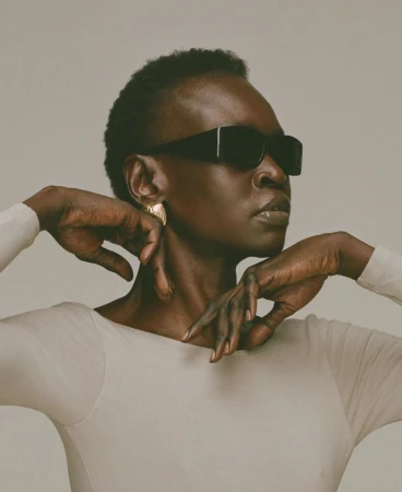 Dark-skinned woman with short hair wearing sunglasses and earrings, posing in a light-colored long-sleeved shirt in a photo studio.