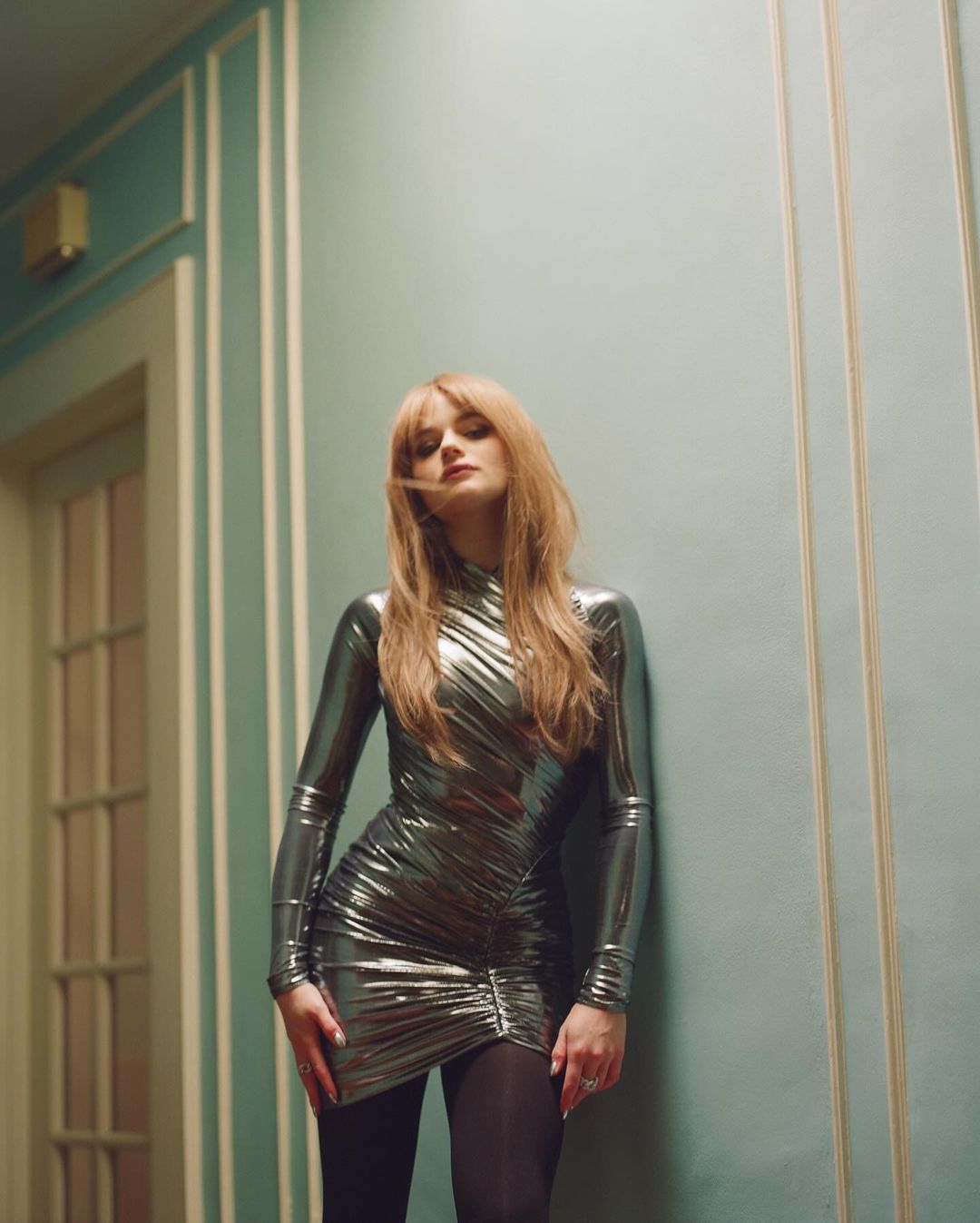 Elegant woman in a metallic silver dress poses indoors against a teal wall. She exudes sophistication in a high-fashion style.