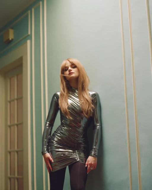 Elegant woman in a metallic silver dress poses indoors against a teal wall. She exudes sophistication in a high-fashion style.
