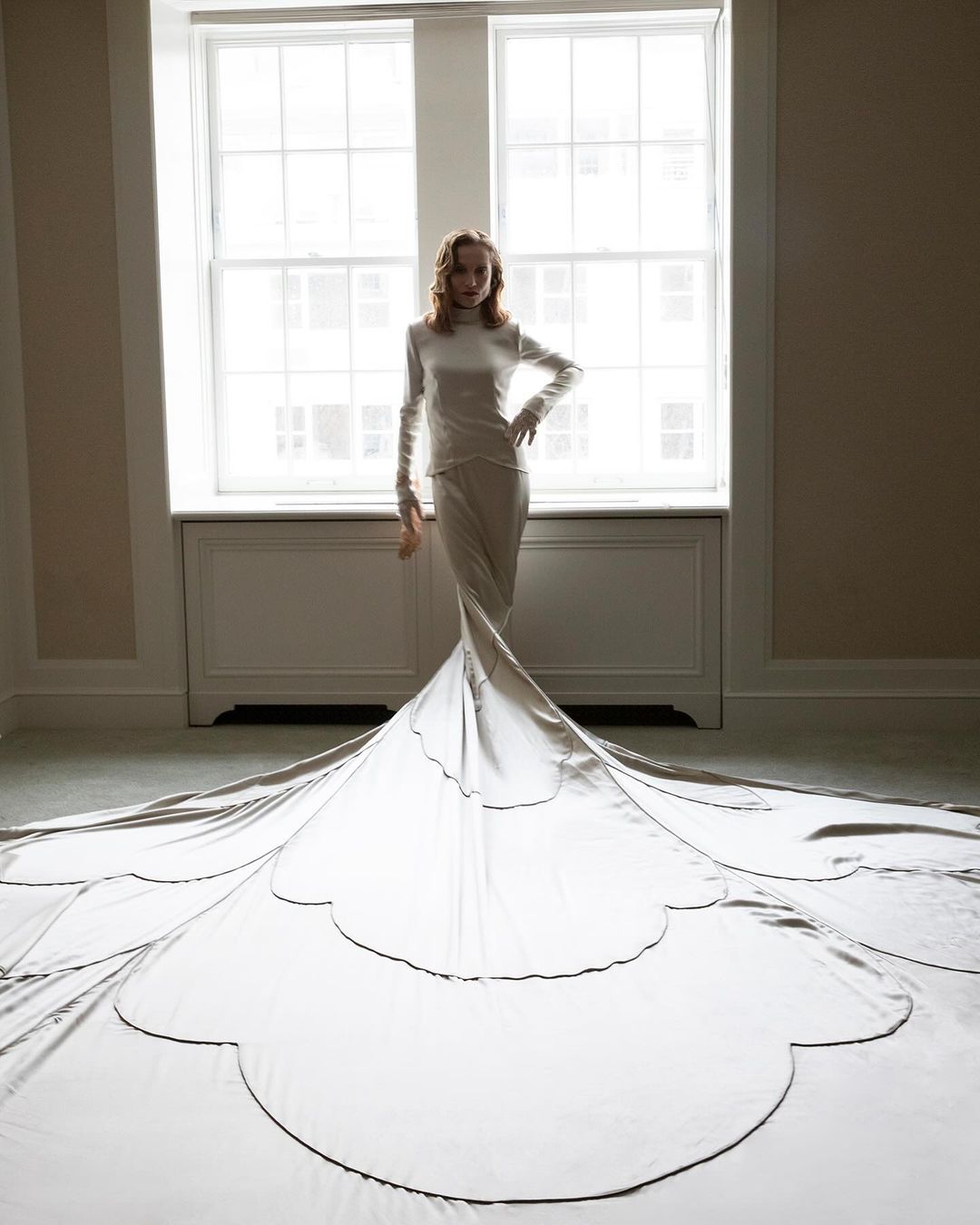 Full-length shot of a woman in a long-sleeve dress with a large train, posing in front of a window in a room with neutral walls.