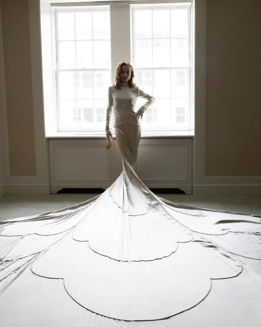 Full-length shot of a woman in a long-sleeve dress with a large train, posing in front of a window in a room with neutral walls.