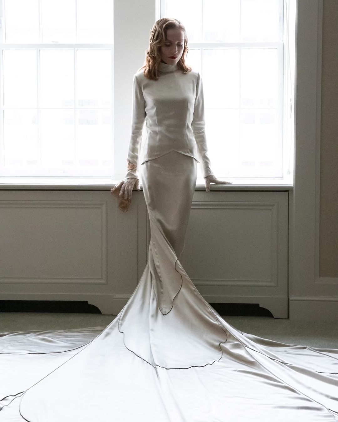 Adult female model poses in a satin gown with a long train in front of a window with natural light, wearing matching gloves.