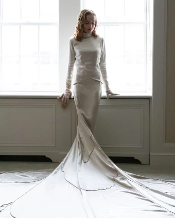 Adult female model poses in a satin gown with a long train in front of a window with natural light, wearing matching gloves.