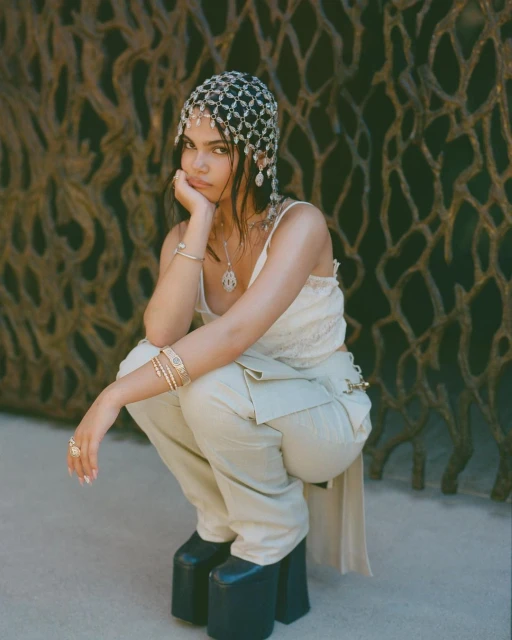 Woman poses in a lace top, trousers, and platform shoes, adorned with jewelry and a beaded headpiece, in front of a textured backdrop.
