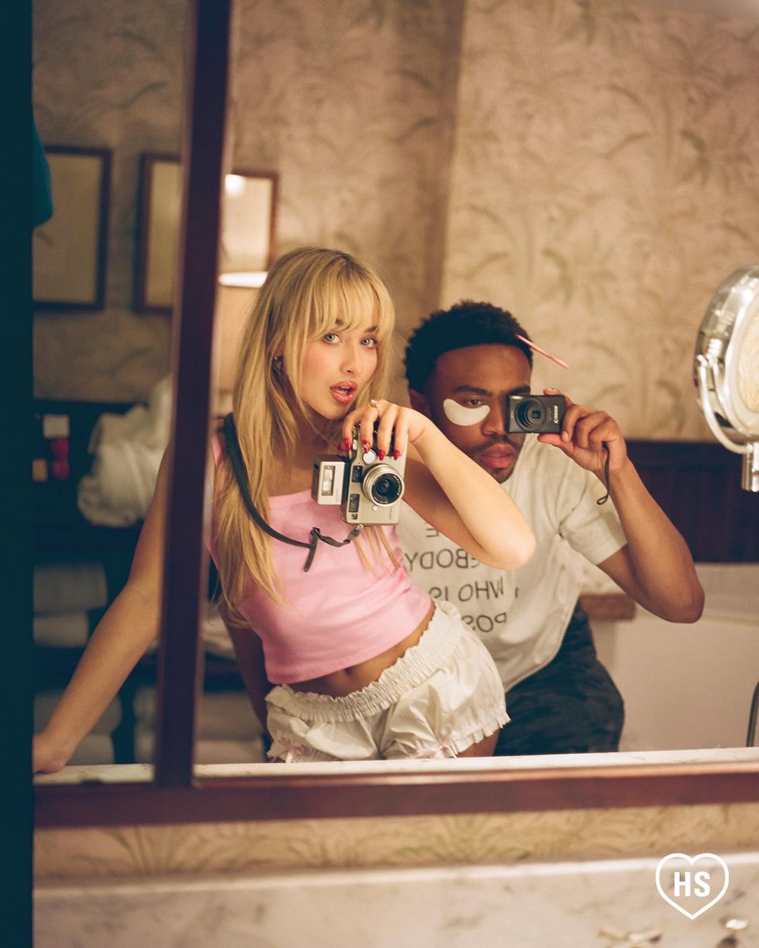 Young woman and man taking a selfie in a mirror with vintage cameras, wearing casual outfits in an indoor setting with wallpaper.