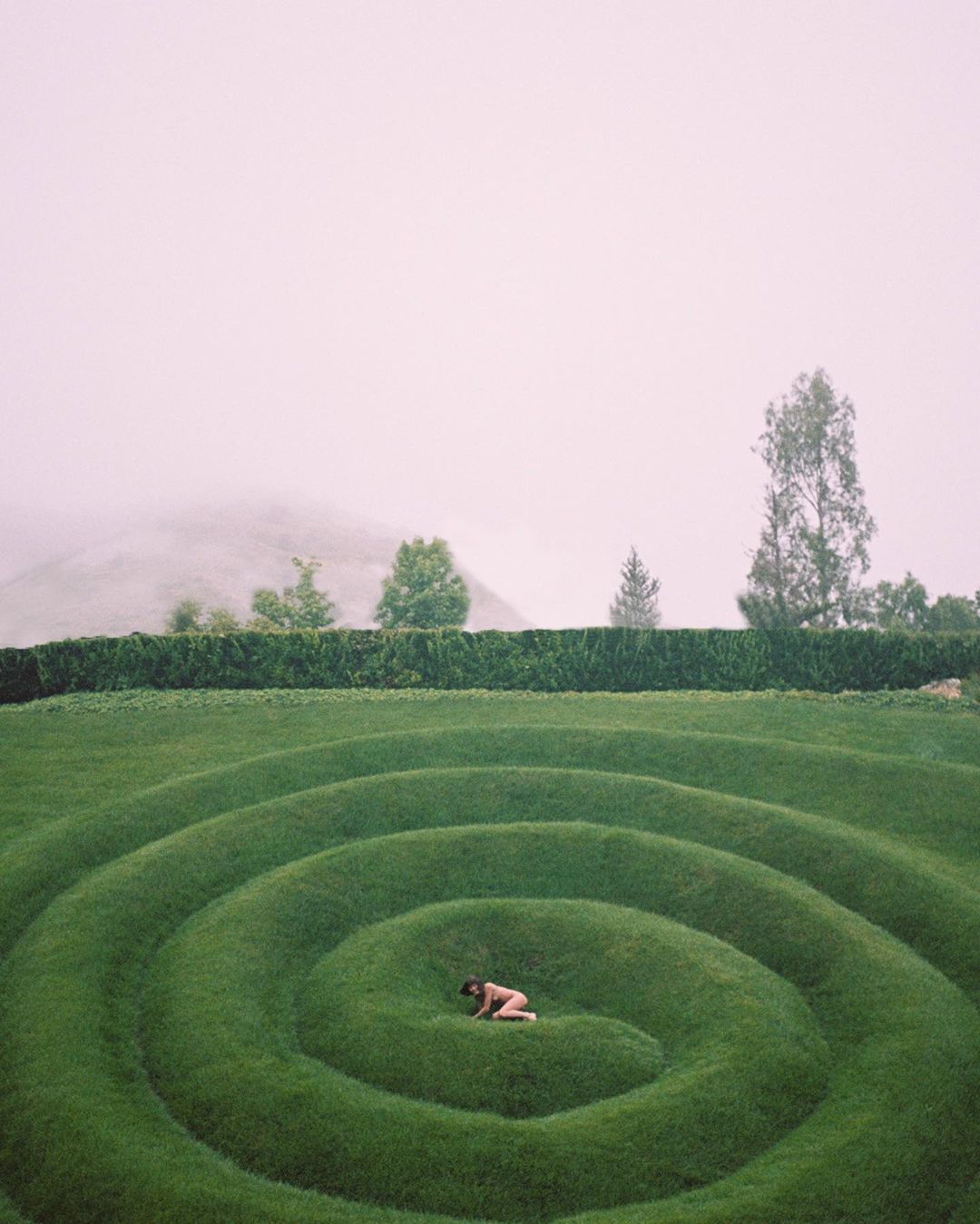Person lies on a circular grass maze on an overcast day. Green grass, trees, hedge, and distant hill with soft, diffused lighting.