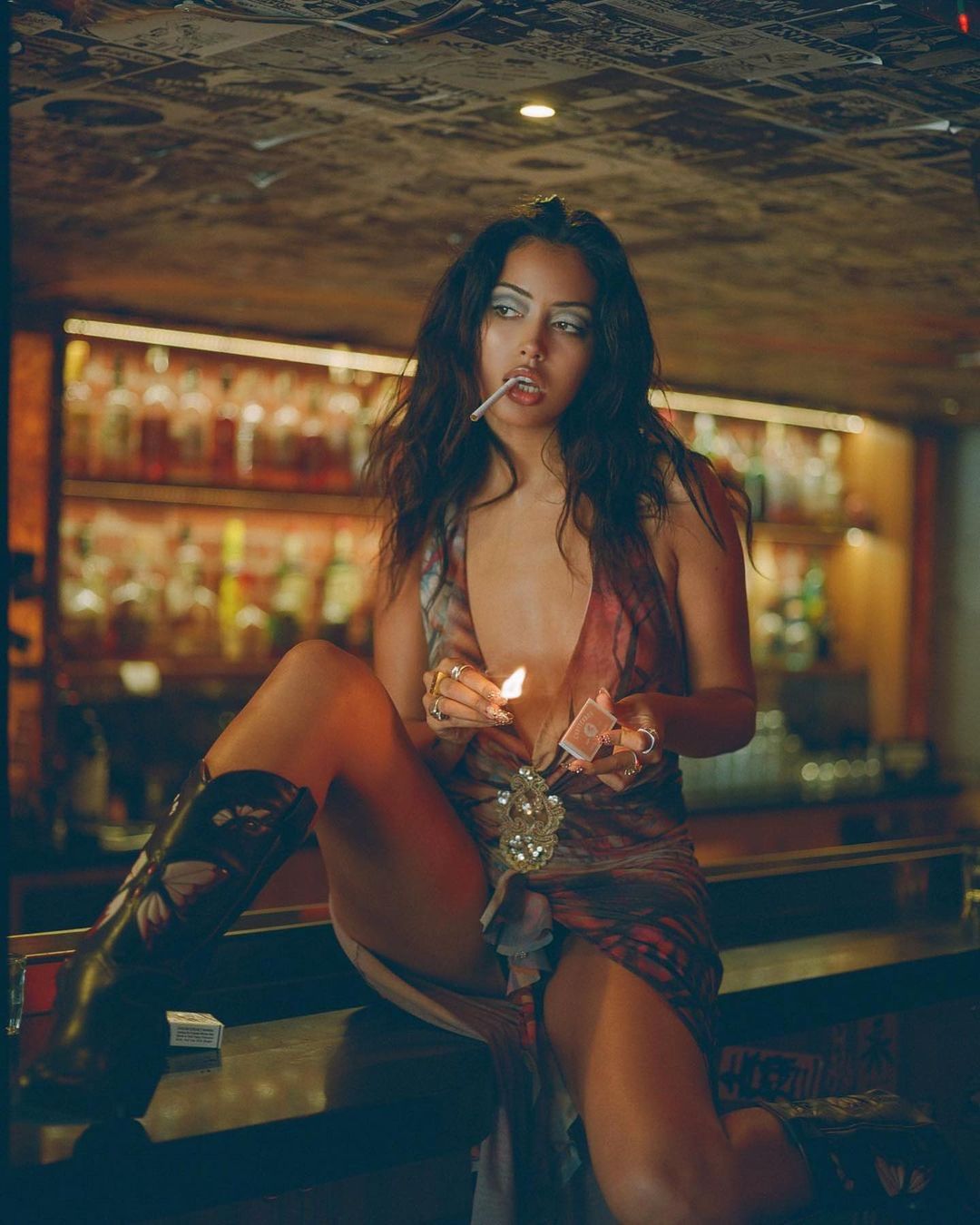 Woman in patterned dress sits on a bar, smoking and lighting a match. Bottled liquor displayed on shelves in the background.