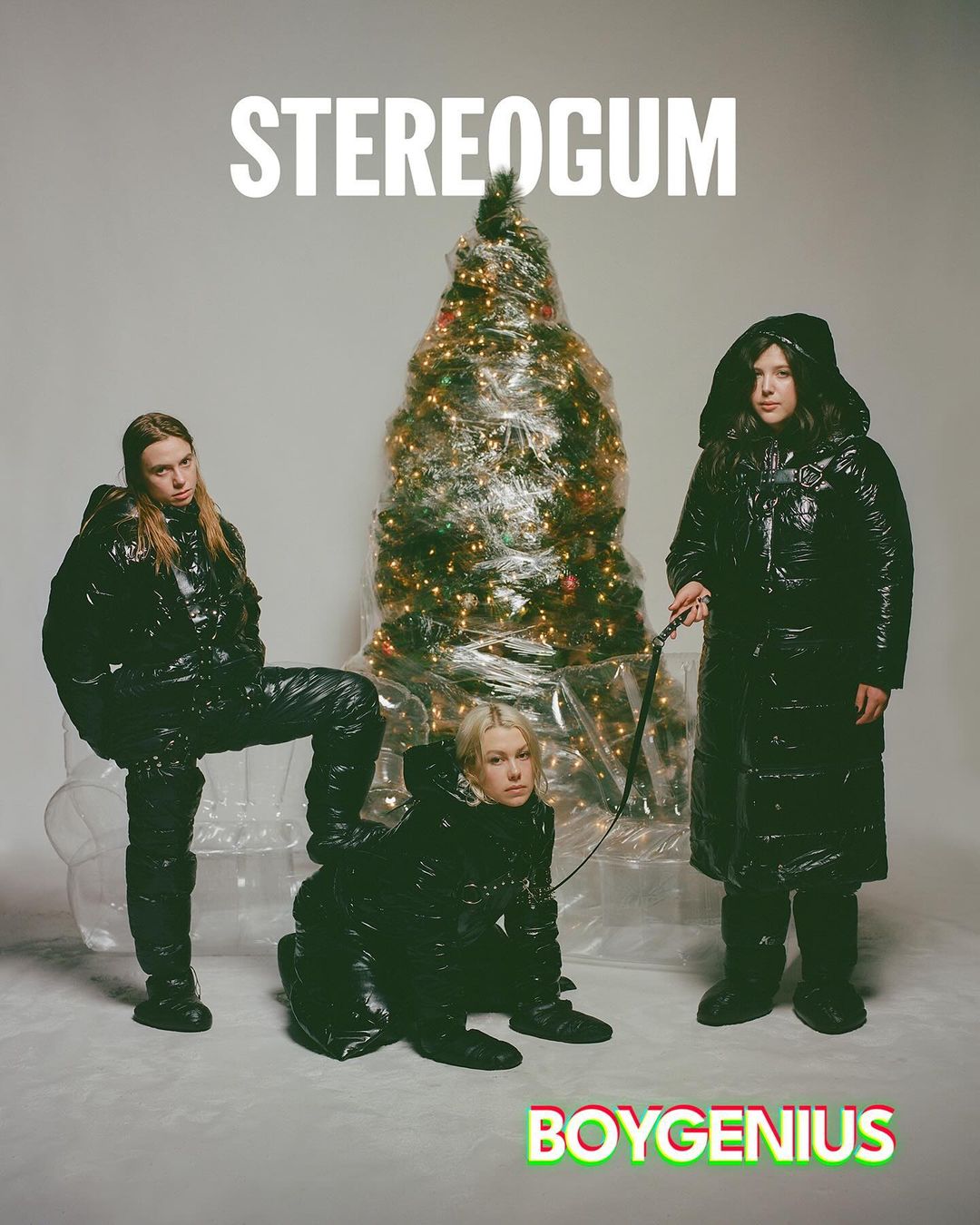 Three individuals pose in shiny black puffer coats alongside a Christmas tree wrapped in plastic. One woman kneels while being leashed.