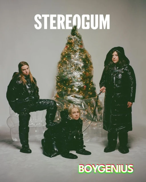 Three individuals pose in shiny black puffer coats alongside a Christmas tree wrapped in plastic. One woman kneels while being leashed.
