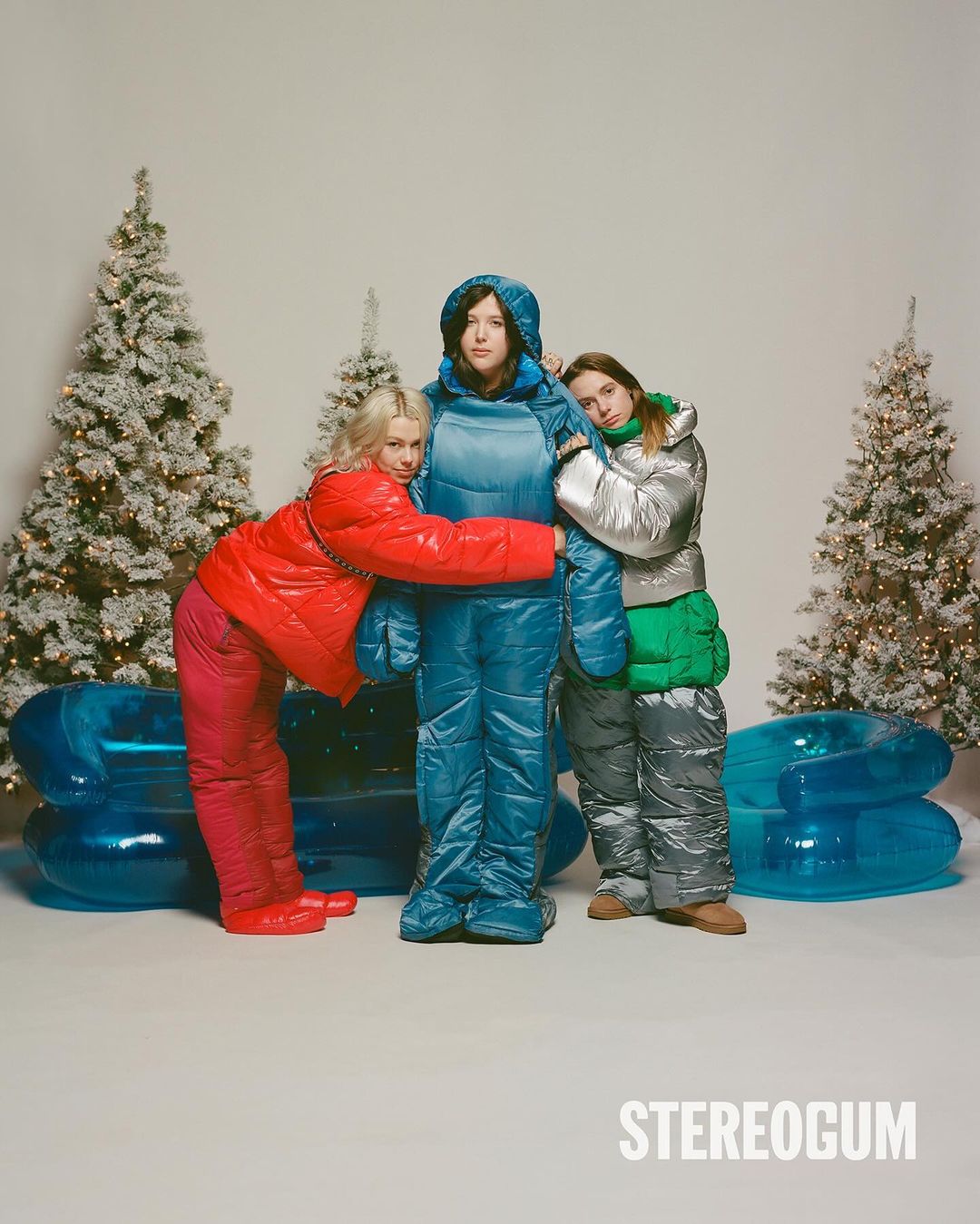 Three women in insulated outerwear standing near decorated Christmas trees and an inflatable couch in a festive, wintry setting.