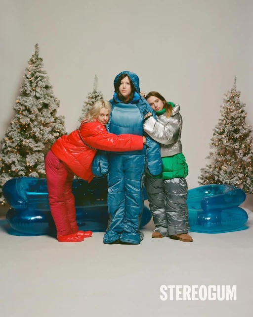 Three women in insulated outerwear standing near decorated Christmas trees and an inflatable couch in a festive, wintry setting.
