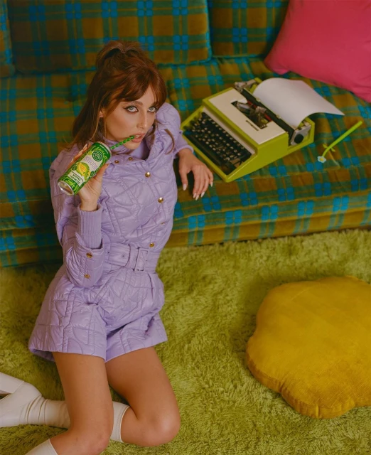 A woman with red hair in a purple outfit holds a lemon-lime can while kneeling on a green carpet with a retro couch and typewriter.