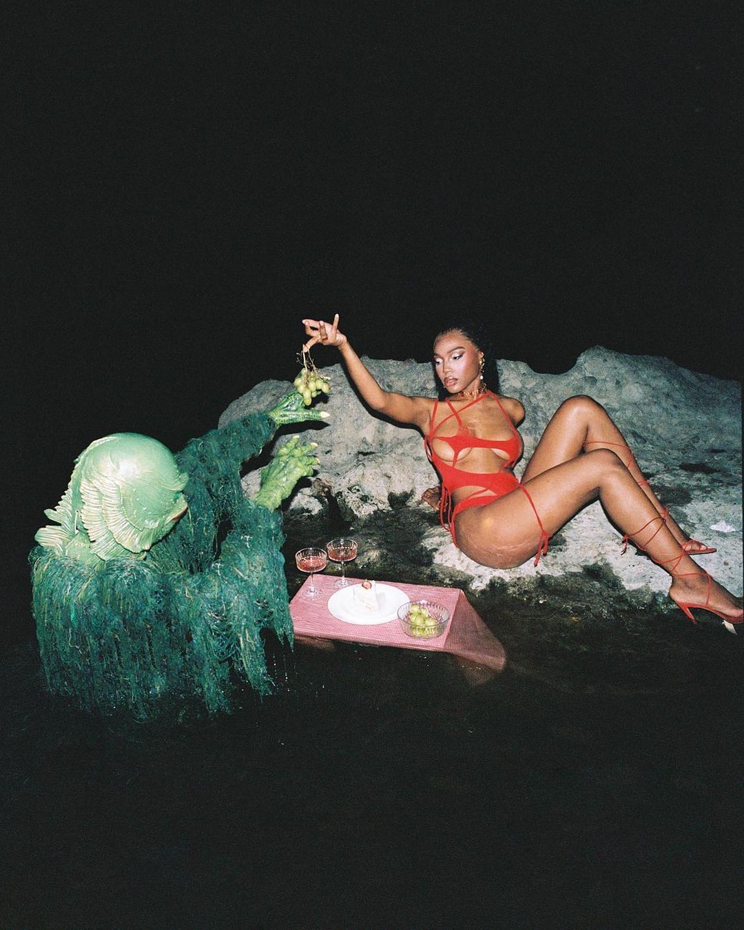 Woman in a red bathing suit sits beside a swamp creature at a dark waterside picnic with grapes, wine, and olives.