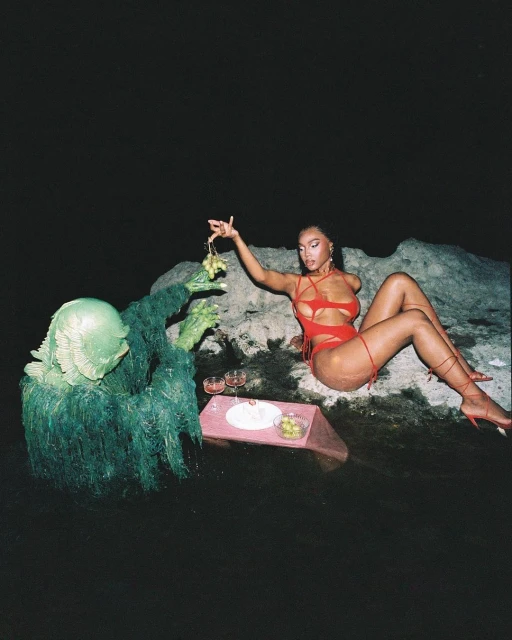 Woman in a red bathing suit sits beside a swamp creature at a dark waterside picnic with grapes, wine, and olives.