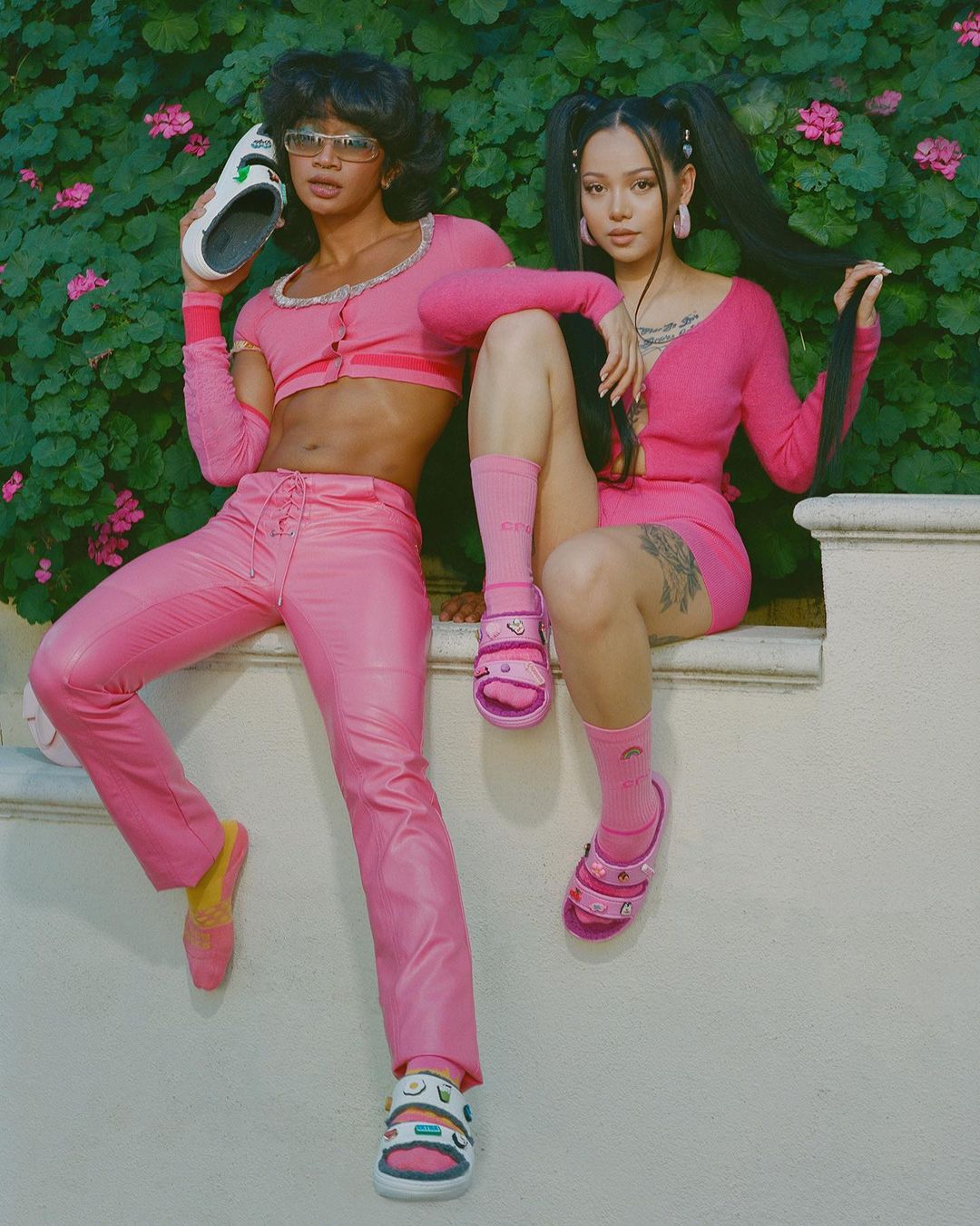 Two women pose on a ledge wearing pink outfits and pink and white shoes with a floral background. One holds a shoe.