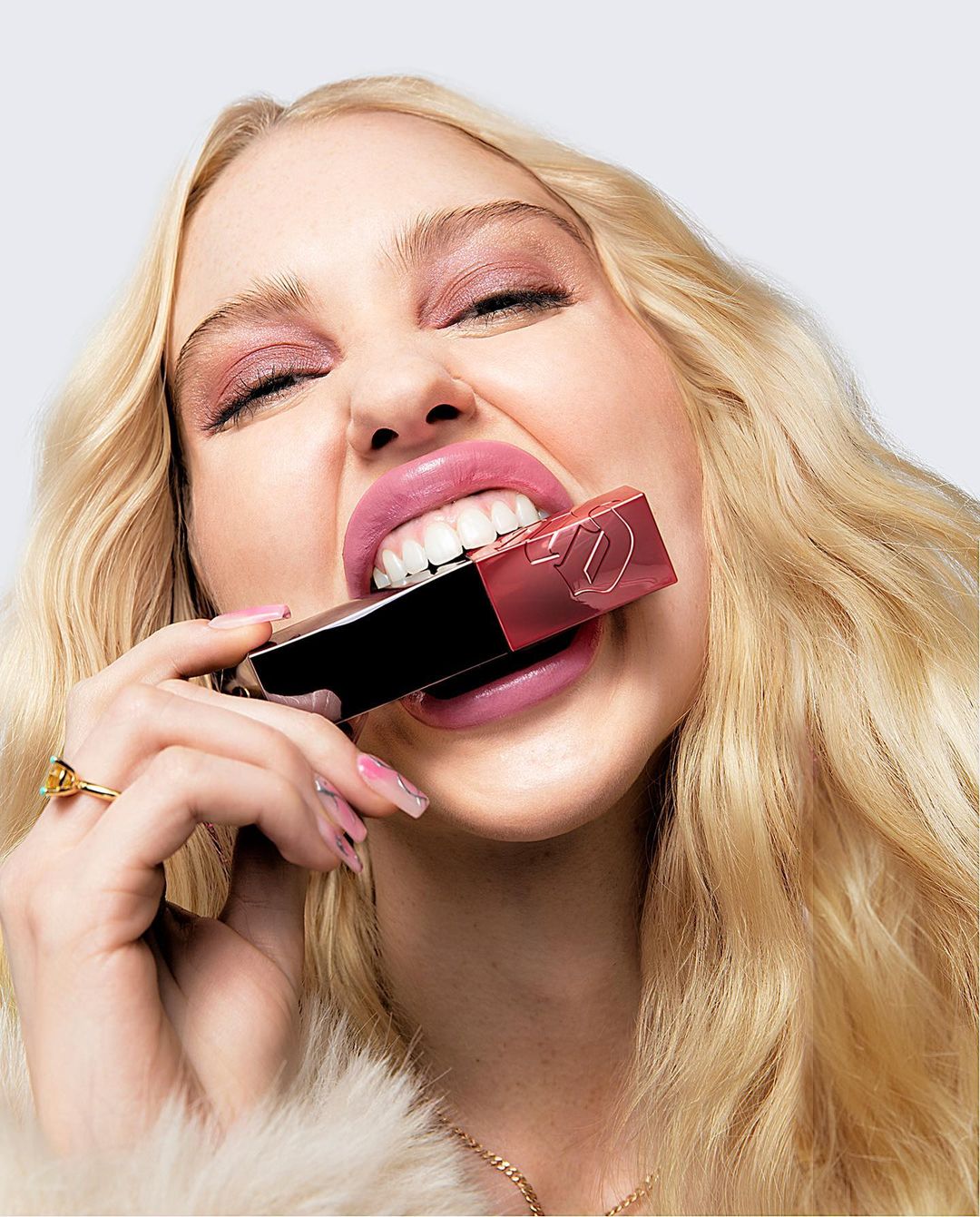 Blonde woman playfully bites a tube of lipstick, showcasing a beauty campaign with focus on makeup and bright studio lighting.
