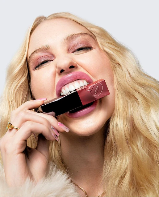 Blonde woman playfully bites a tube of lipstick, showcasing a beauty campaign with focus on makeup and bright studio lighting.