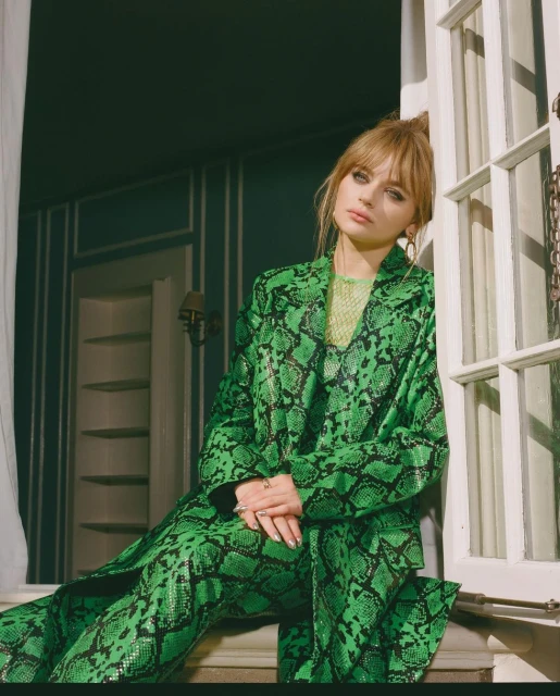 A woman in a green snakeskin suit poses in front of a window. She has blonde hair and is looking at the camera.