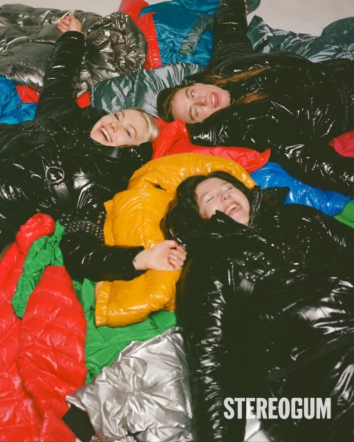 Three adult women lie on a colorful collection of glossy puffer jackets indoors, smiling and laughing, with a high-angle, eye-level perspective.