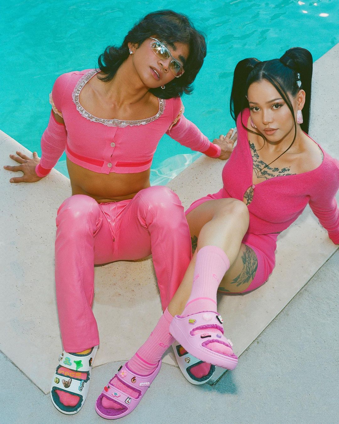 Two stylish people, a male and a female, pose poolside wearing pink outfits and colorful Crocs, capturing a fashion-forward, summer vibe.