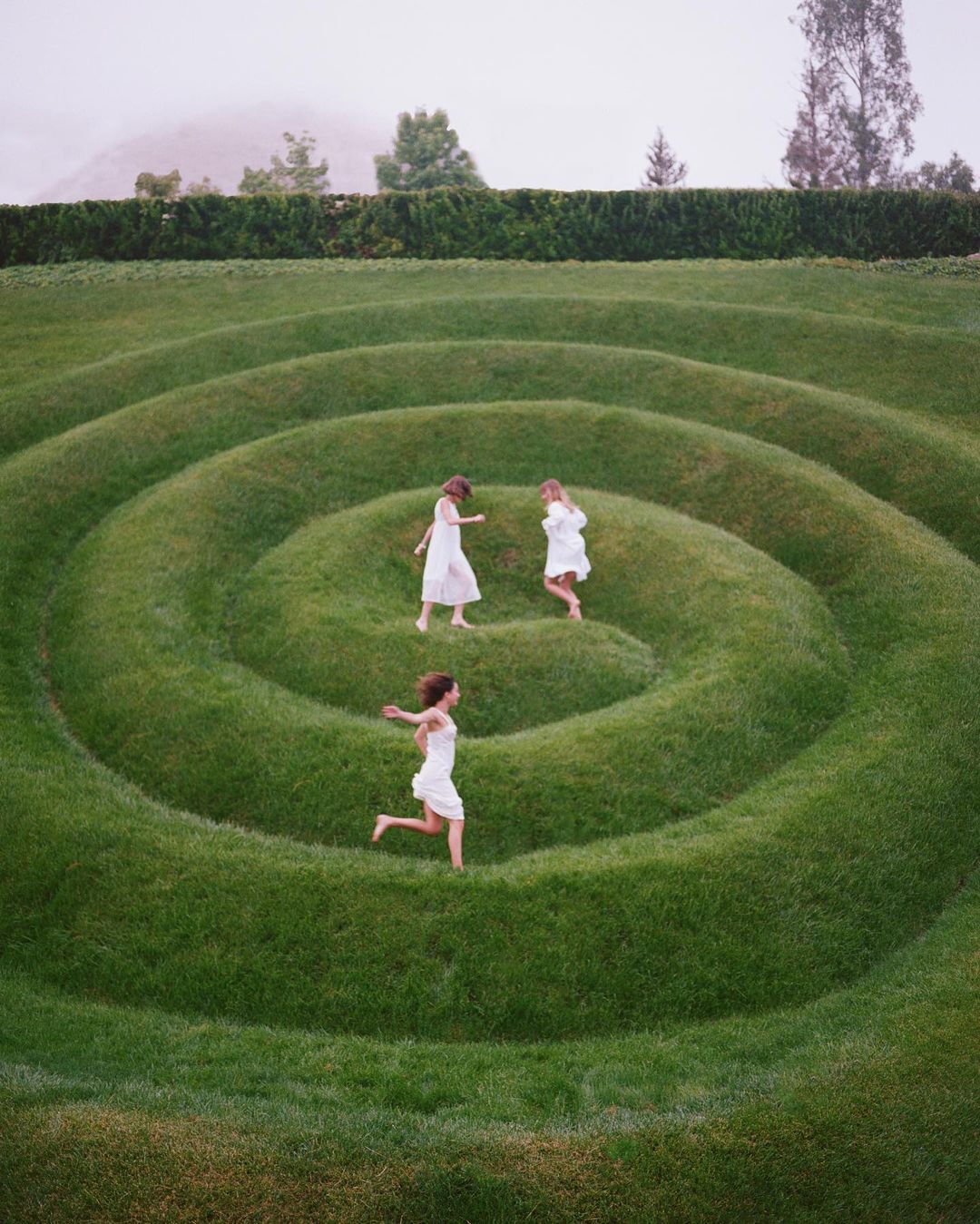 Three children in white dresses play and run through a spiral grass garden on a cloudy day in a wide, eye-level shot.