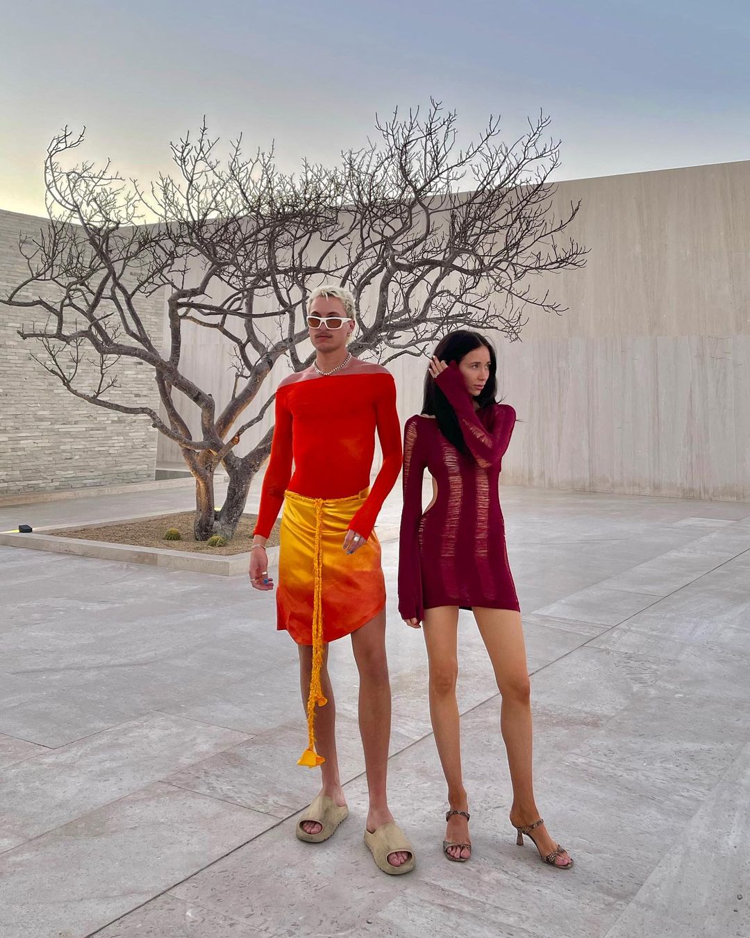 Two people pose outdoors. One wears a red top and orange skirt, the other a burgundy dress, against a neutral wall and sky.