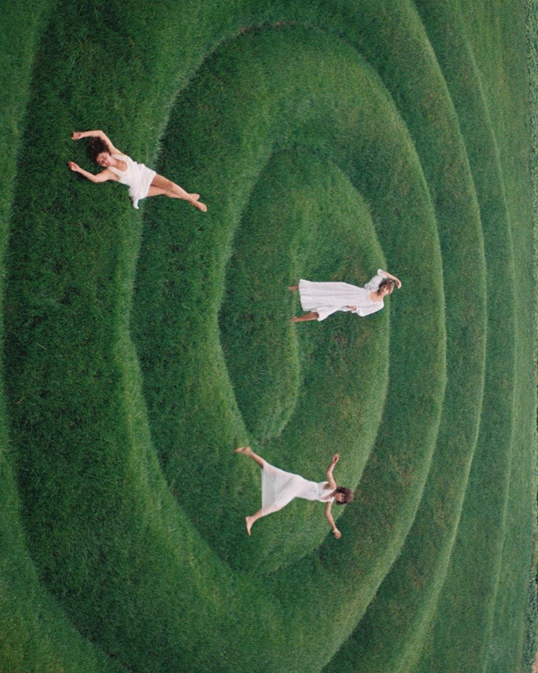Aerial view of three adult women in white dresses lying on a spiral-shaped, terraced, green grass landscape, creating a visually striking composition.