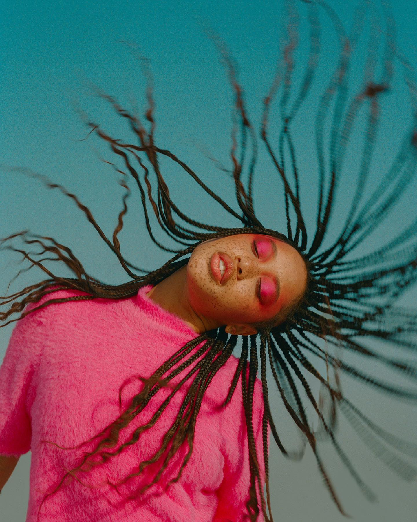 A young adult woman with pink eyeshadow and braided hair poses against a blue sky, wearing a pink sweater, with a dynamic upward tilt.