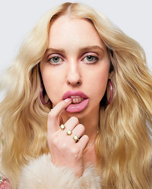 Close-up portrait of a young woman with blonde hair and pink lipstick wearing rings and pink earrings in a photo studio.