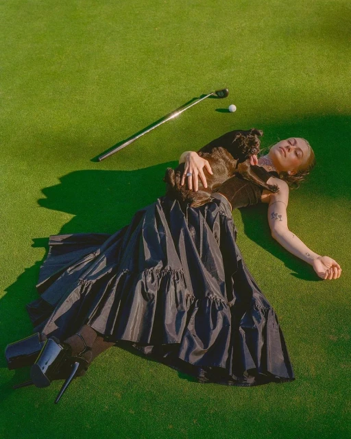Woman with dog on golf course with club and ball, wearing a dark dress and high heels. Lying on green grass in natural light.