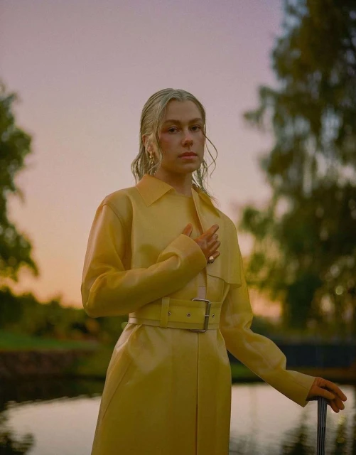 Portrait of a blonde woman in a yellow coat standing near water with trees. Golden hour lighting with soft focus and natural setting.