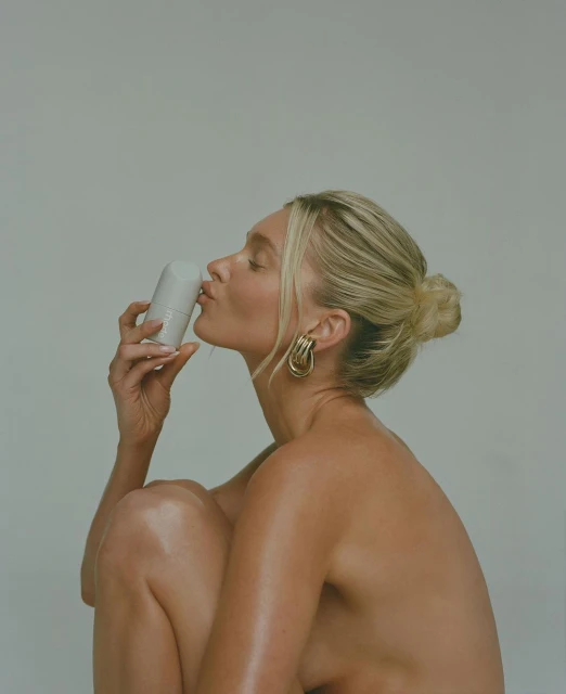 Blonde woman with nude shoulders kisses white deodorant tube in a clean photo studio setting with simple earrings and a hair bun.