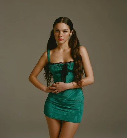 Portrait of a young woman with pigtails wearing a shiny green minidress against a neutral grey studio background.