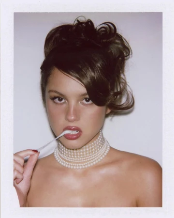Close-up portrait of a woman wearing layered pearl necklaces holding a strand of white chewing gum to her mouth, against a white seamless background.