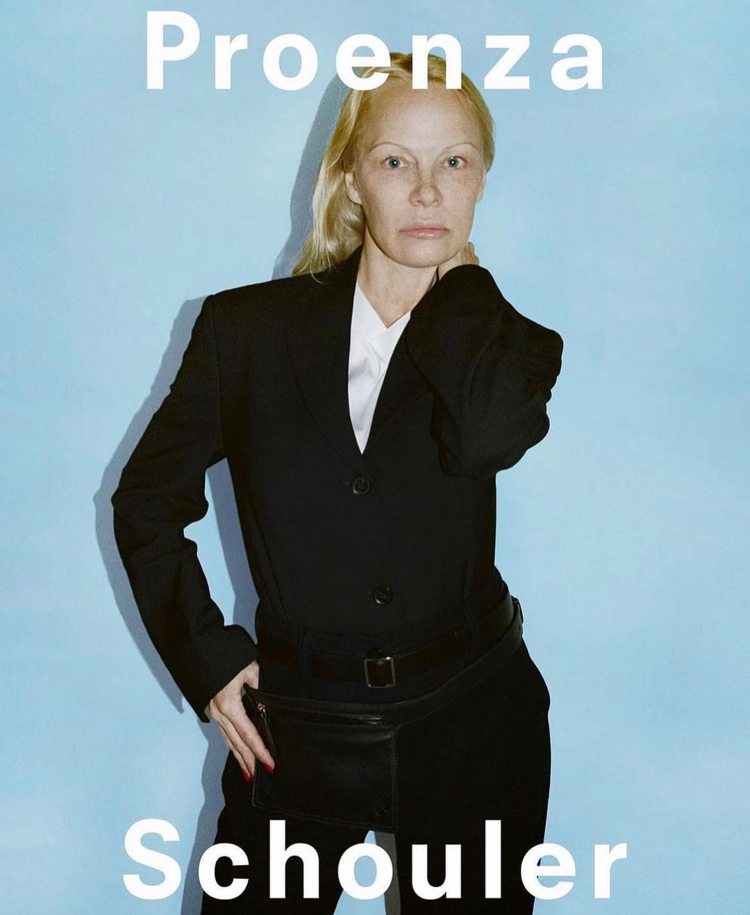 Woman in a black suit and fanny pack poses against a light blue backdrop. Text reads 'Proenza Schouler'. Fashion editorial shot.