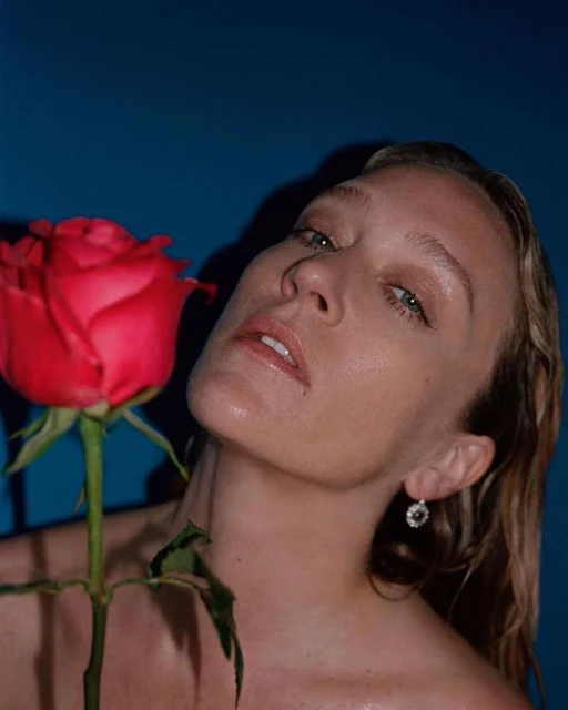 Adult woman with blond hair poses in front of a blue background, holding a red rose. Studio lighting enhances her features.