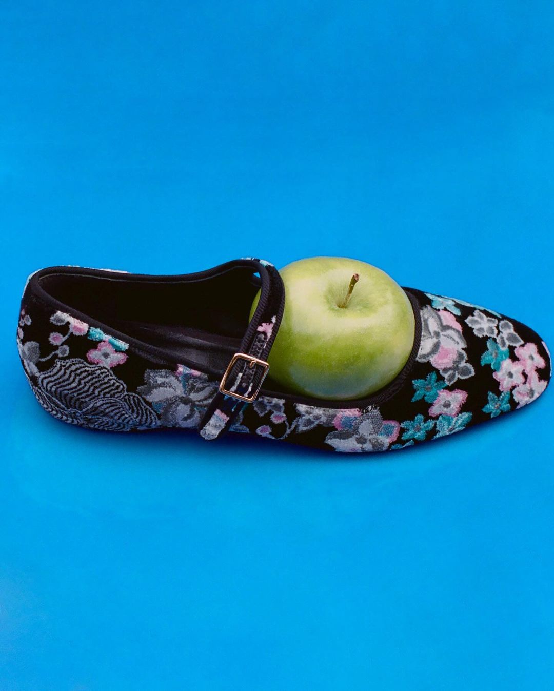 Floral pattern Mary Jane shoe with gold buckle and green apple inside against a vibrant blue backdrop. Close-up still life fashion image.