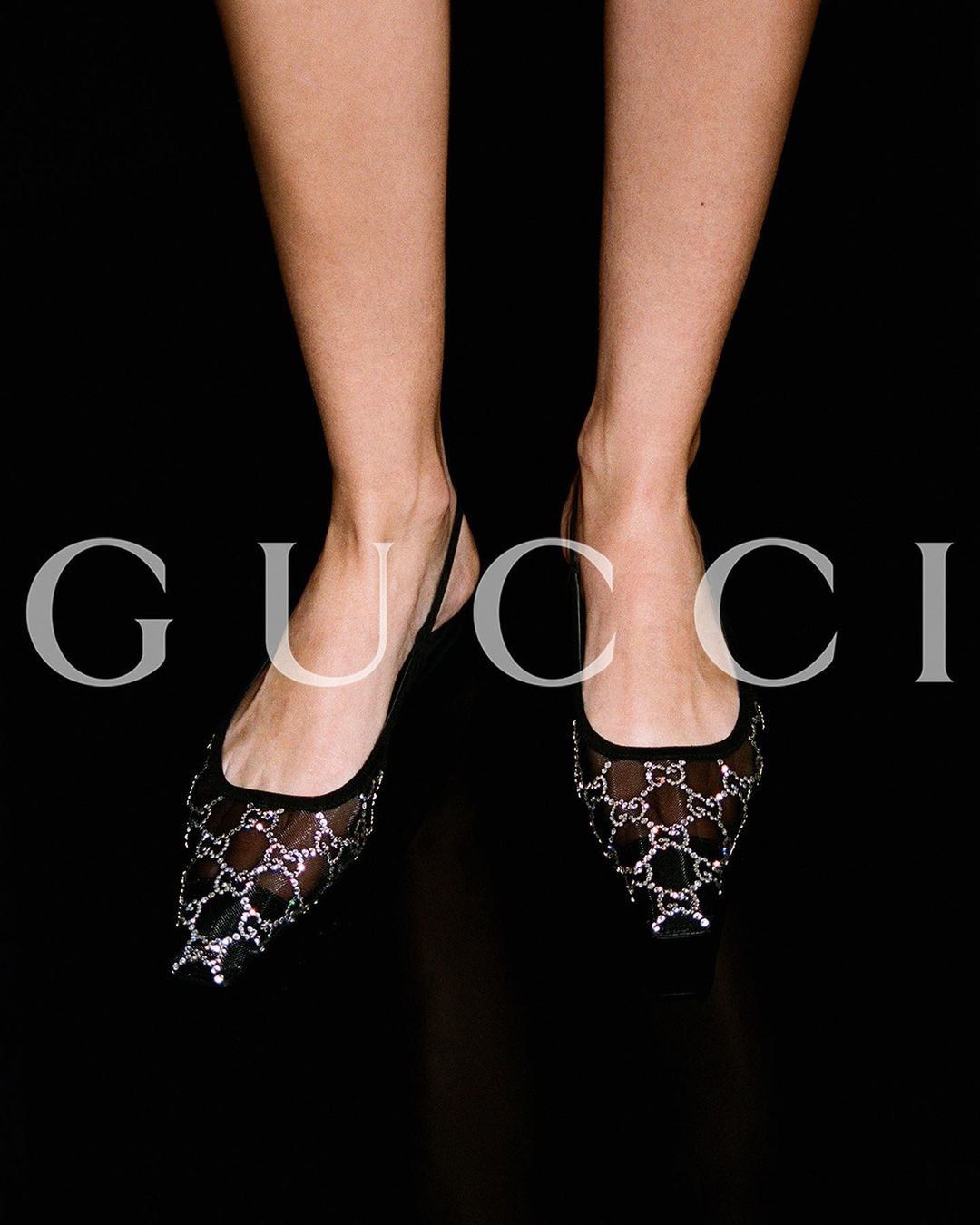 Close-up of a woman's legs and feet wearing black mesh Gucci flats, set against a stark black background highlighting the shoe design.
