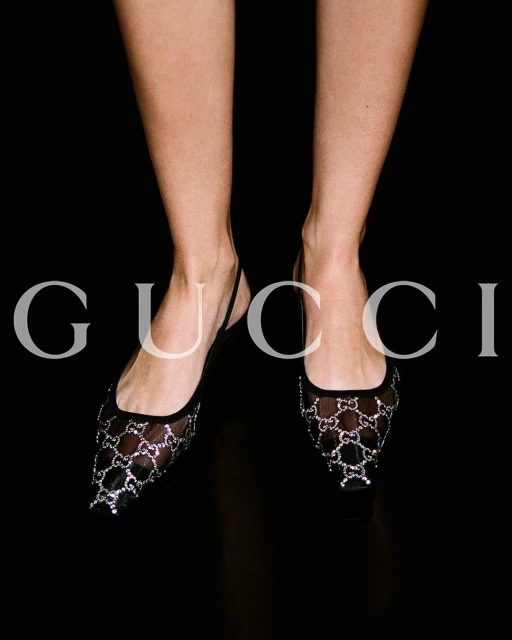 Close-up of a woman's legs and feet wearing black mesh Gucci flats, set against a stark black background highlighting the shoe design.