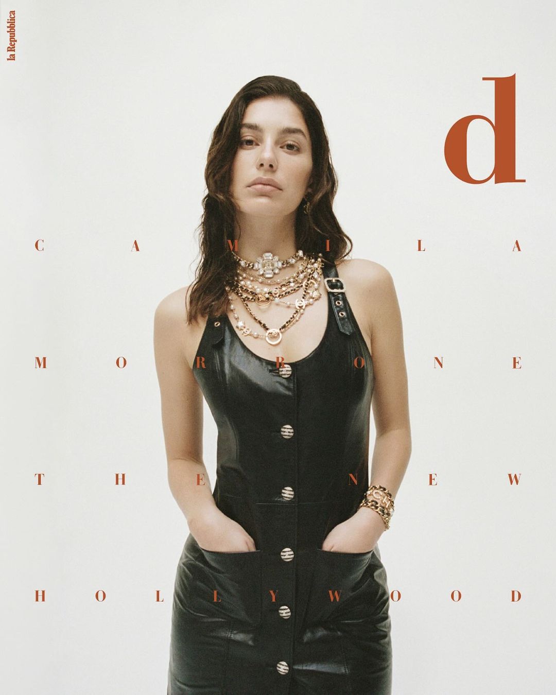 Editorial fashion shot of a woman in a black dress with hands in her pockets, wearing multiple necklaces, against a plain background.